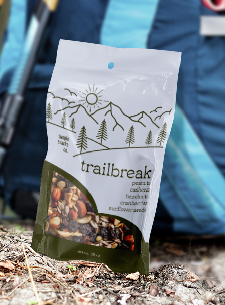 A stand-up pouch labeled Simple Snacks Co. Trailbreak with a line-art mountain and forest design, listing peanuts, cashews, hazelnuts, cranberries, and sunflower seeds, propped upright on gravel against a blurred blue backpack.
